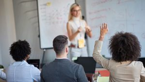 Claim Your Seat - 8 Reasons To Speak Up In The Workplace - a group of people in a work presentation with one person raising their hand.